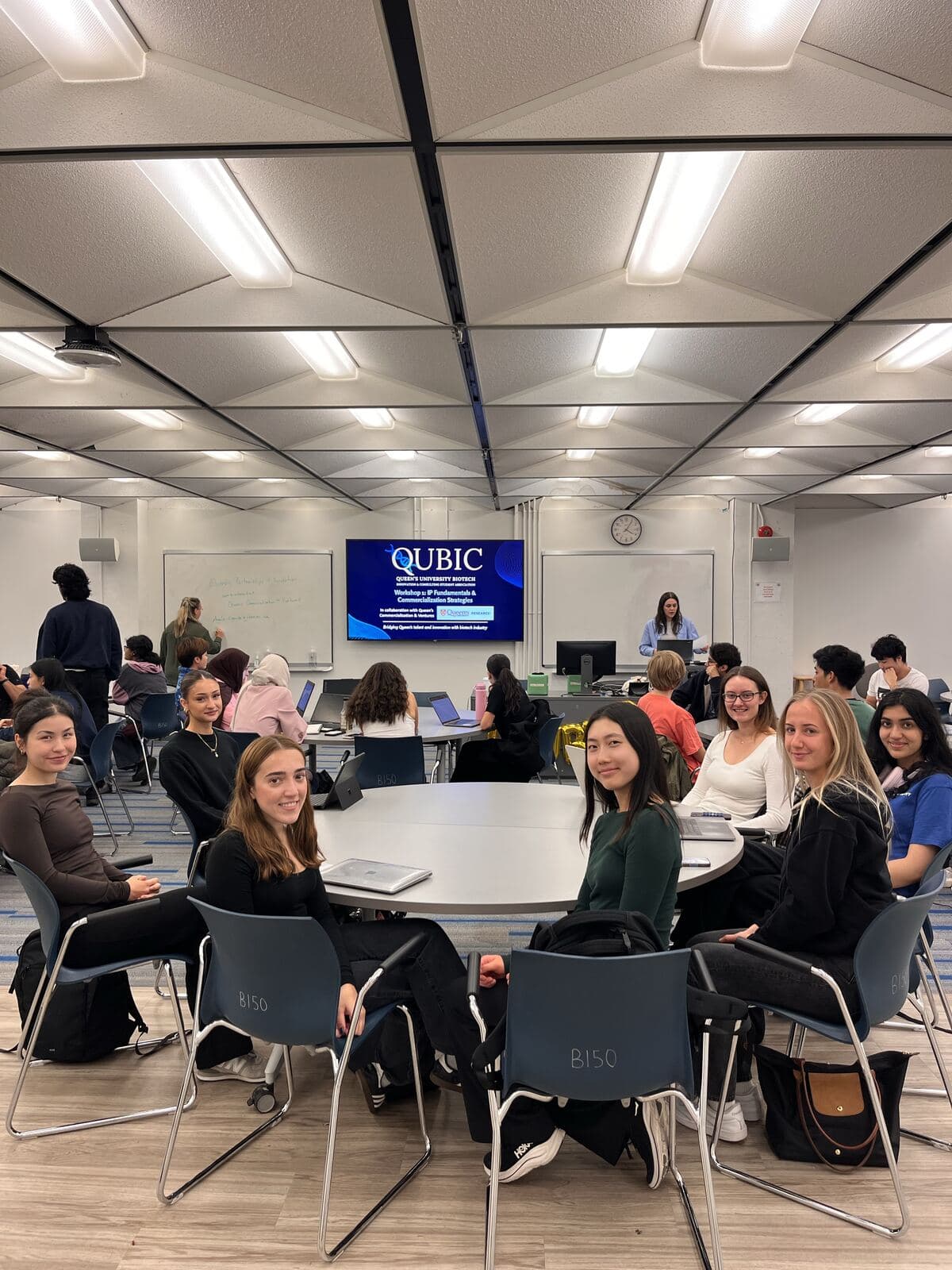 Students seated at a round table smiling at the camera, with the QUBIC Workshop 1 title slide on the screen behind them