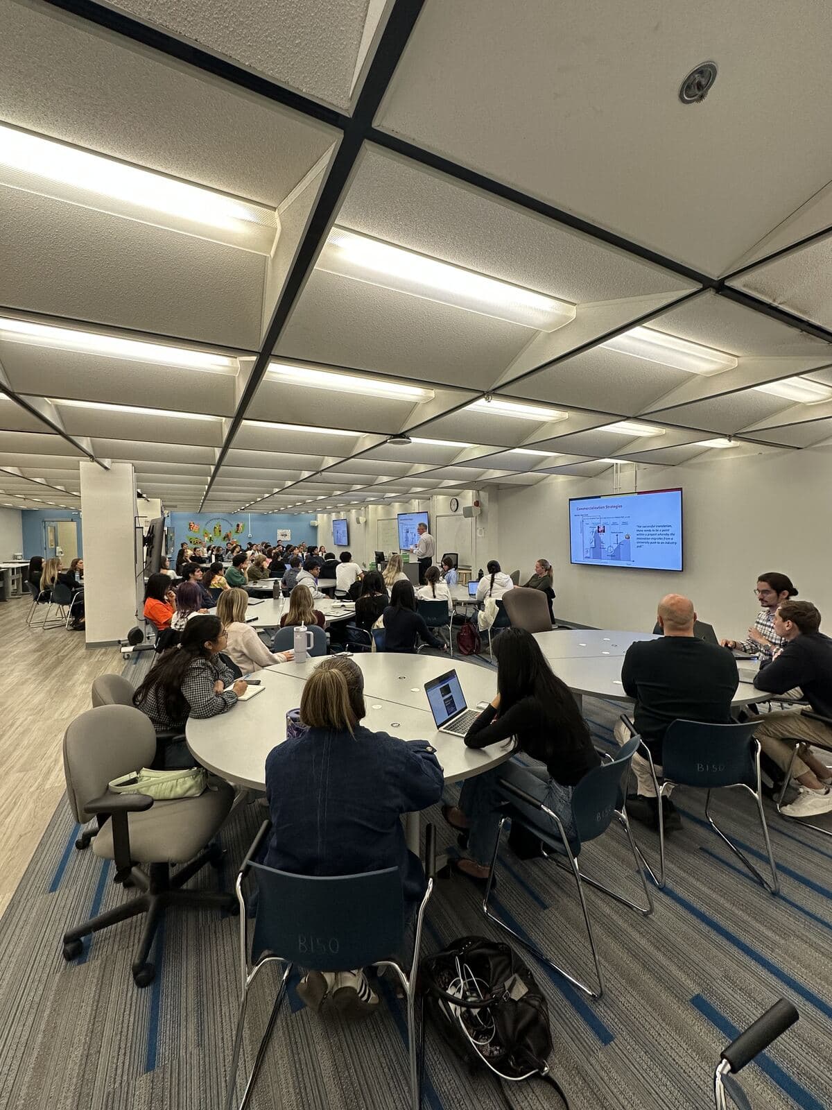 A wide view of the full workshop room, with students at round tables facing the speaker